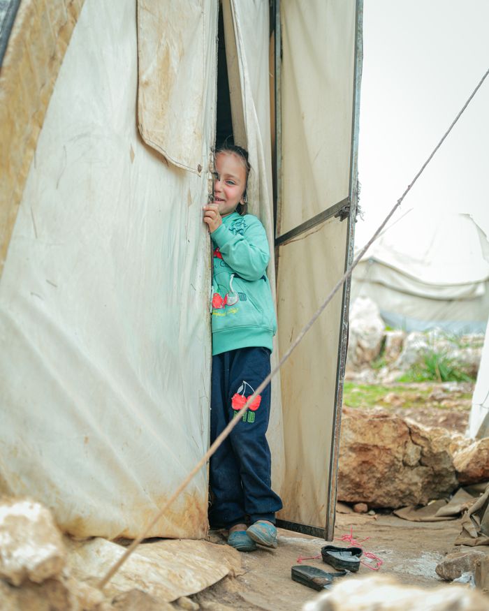 about-us A young girl with blonde hair peeks out from a tent in a Syrian refugee camp, capturing innocence and resilience.