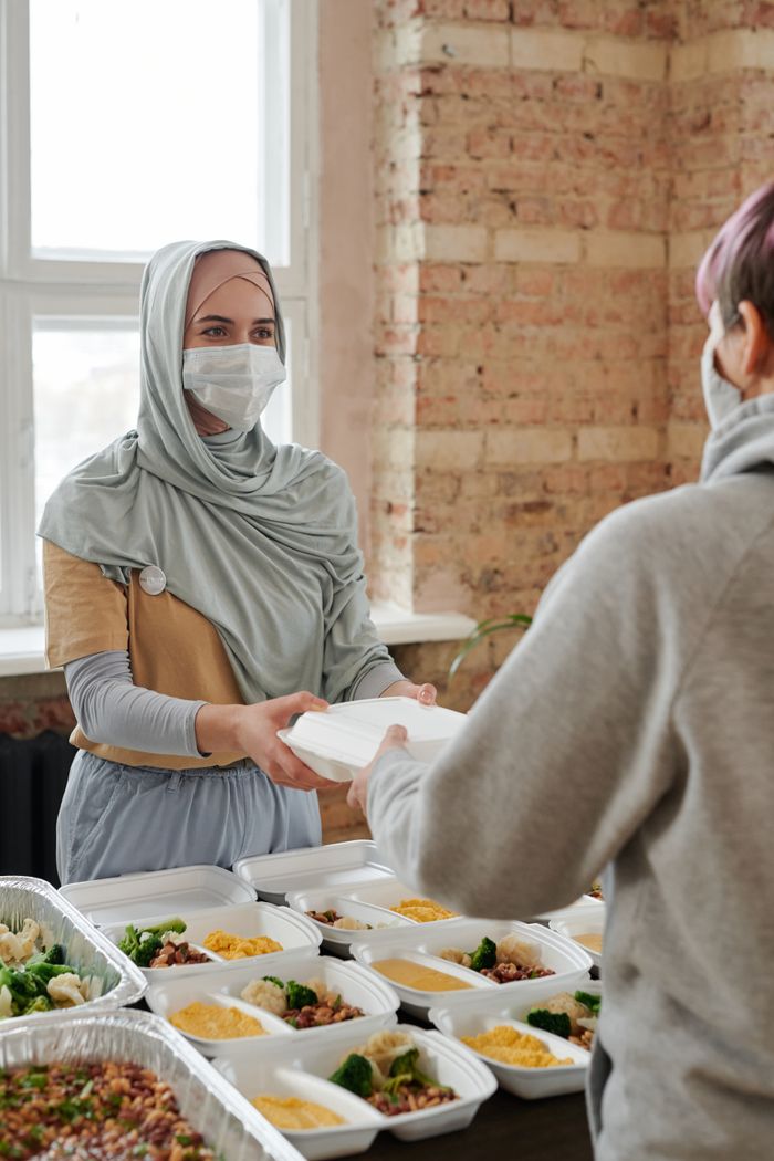 Mastering the First Impression: Your intriguing post title goes here A woman wearing a hijab and face mask hands out packed food indoors, promoting charity and safety.
